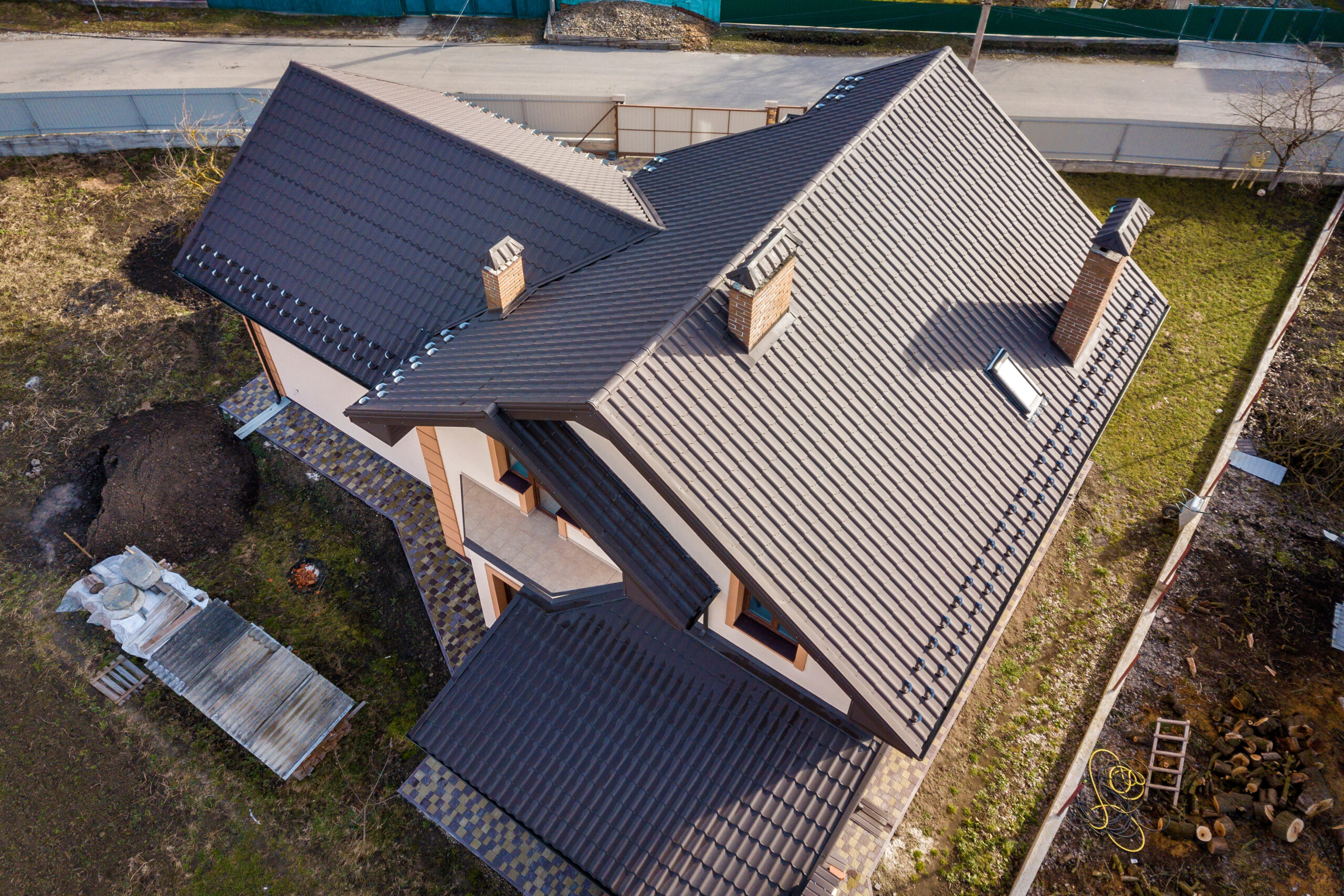 Aerial view of a modern house with a dark metal tile roof and several brick chimneys, surrounded by a yard and construction materials.