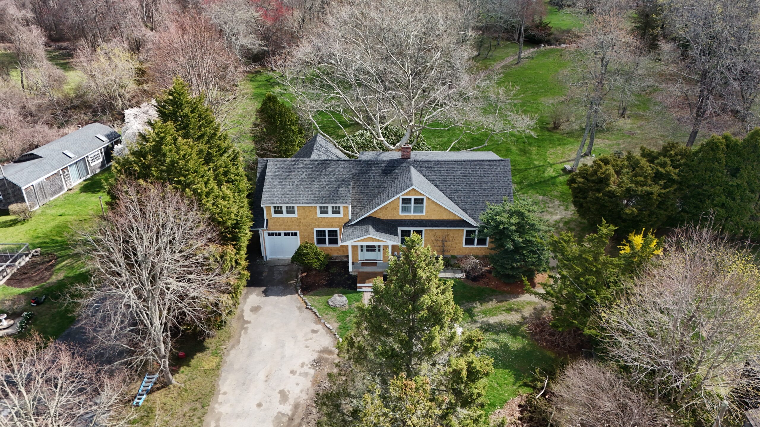 Aerial view of a yellow two-story house with a gray roof, white trim, and a long driveway surrounded by bare trees and green lawn.